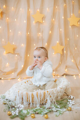 Fototapeta premium Little cute girl 1 year old in a light knitted sweater and bandage with a flower on her head sits in a basket decorated with needles and Christmas balls. Festive mood. Merry Cristmas