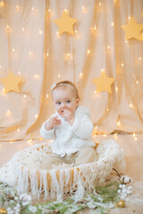 Fototapeta premium Little cute girl 1 year old in a light knitted sweater and bandage with a flower on her head sits in a basket decorated with needles and Christmas balls. Festive mood. Merry Cristmas