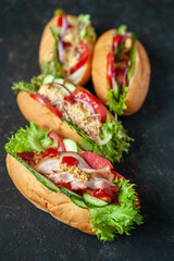 Four hot dogs on a dark background. Fresh tomatoes, cucumbers, lettuce, sausage, bacon, cheese, mustard and ketchup.
