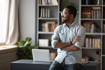 Happy confident young african businessman freelancer standing near workplace desk in modern office...