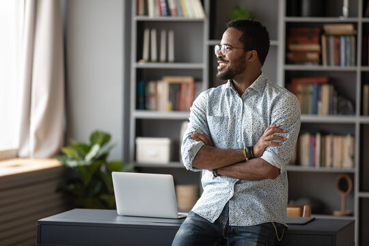 Happy confident young african businessman freelancer standing near workplace desk in modern office with arms crossed look out the window dreaming about new career opportunities and great future vision