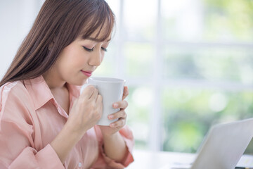 Beautiful Asian woman with long hair Wearing a pink shirt standing, holding a white coffee cup and smell the coffee in a cup. Drink coffee, coffee shop, coffee break concept.