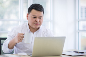 Asian Men Wearing a white shirt holding a white coffee cup and look at the laptop. Relaxing and drinking coffee in office, coffee shop, coffee break concept.