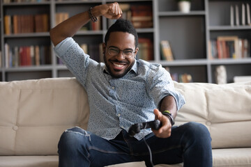 Excited successful african guy sit on couch in living room spend weekend free time at home holding joypad celebrating victory. Having fun using modern technology devices virtual video games concept