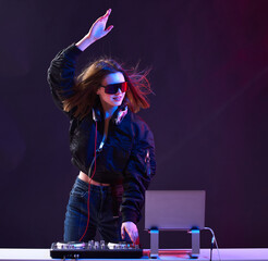 Stylish DJ girl with headphones and glasses on the bomber jacket mixes the music at the party and...