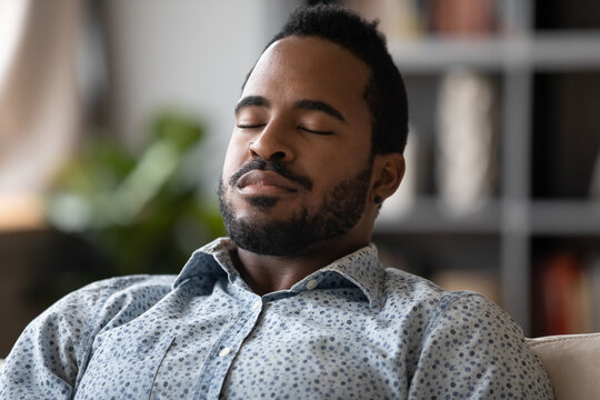 Close Up African Young Man Sit On Couch Closed Eyes Resting Reduces Fatigue, Enjoy Lazy Day, Breath Fresh Air At Conditioned Home, Visualizing And Meditating Improve Inner Balance, No Stress Concept