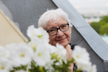 Outdoor close up portrait of beautiful mature wrinkled gray haired woman dressed in stylish blouse,  wearing spectacles, surrounded with flowers, enjoying good wether in summer time.