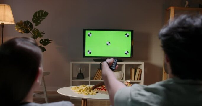Couple spending time together in the evening, sitting on couch and watching tv with green chroma-key green screen. A template for your clips 4k footage