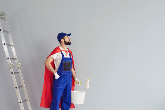 Happy home repair worker engaged in house renovation on gray background. Young contractor holding roller brush, wearing superhero costume near grey wall.