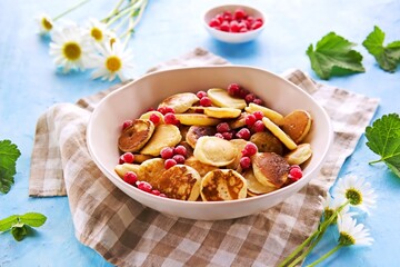 Mini pancakes with red currants in a beige bowl on a blue concrete background.