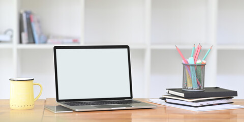 Blank screen computer laptop is putting on a wooden working desk surrounded by office equipment.