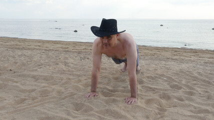 Adult athletic shirtless male in black cowboy hat doing push ups exercising on sandy beach at sunny day