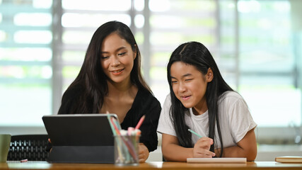 A young mother sits and doing homework together with her daughter at the wooden student desk.