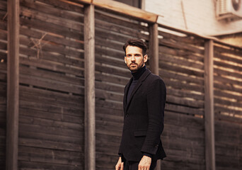 Serious business man in black coat posing near the fence on street autumn background.