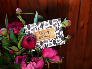 Close-up of pink peonies flowers and vase on wooden background with Happy Birthday card. The photo was taken with natural daylight.