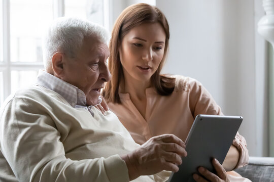 Caring Grownup Daughter Teaching Older Father To Use Computer Tablet Close Up, Pointing At Screen, Mature Man Asking Questions About Mobile Device To Young Woman, Elderly And Technology Concept