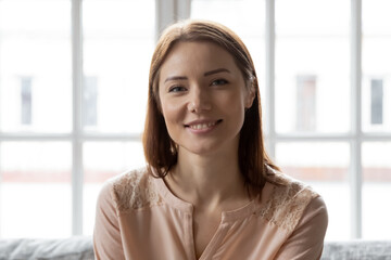 Head shot portrait close up smiling young woman looking at camera, sitting on cozy couch at home, beautiful female making video call, remote job interview, vlogger recording vlog for social network