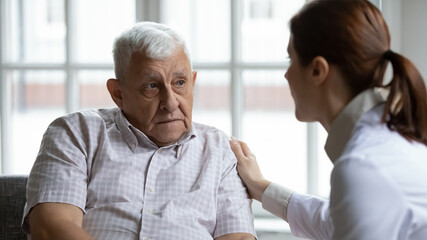 Female doctor comforting upset older patient at meeting, touching shoulder, expressing empathy and support, sad unhappy lonely mature man looking at caregiver close up, elderly healthcare concept