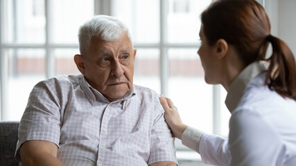 Fototapeta premium Female doctor comforting upset older patient at meeting, touching shoulder, expressing empathy and support, sad unhappy lonely mature man looking at caregiver close up, elderly healthcare concept