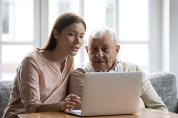 Caring grownup daughter teaching elderly father to use laptop, young woman and mature man looking at computer screen, sitting on couch at home, watching movie, making video call or shopping online