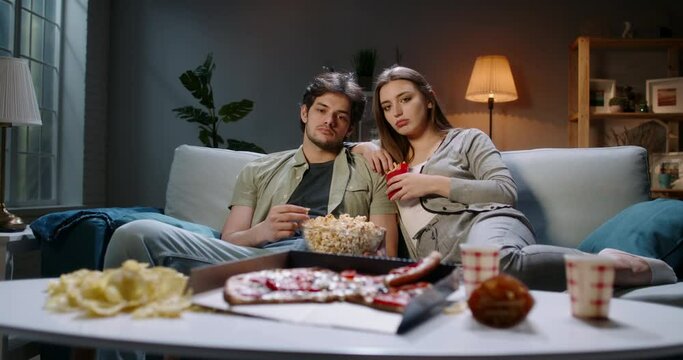 Young student caucasian couple spending time together on couch, sitting in front of tv, watching random shows in absolute boredom, tired after a long day eating unhealthy food 4k footage