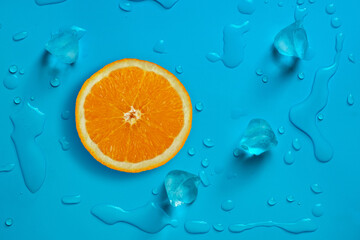 Fresh juicy slice of orange and ice cubes on the bright blue background covered with water drops.