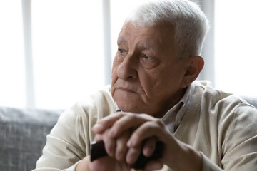 Close up thoughtful sad older man folded hands on wooden cane, walking stick, sitting on couch,...