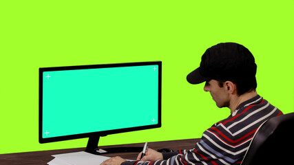 Computer programmer making notes in notebook in front of blank mockup monitor against green screen,...