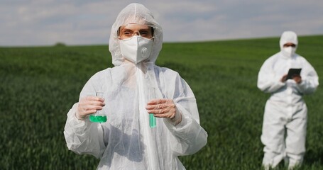 Caucasian woman scientist in protective clothing holding tube with pesticides liquid and doing...