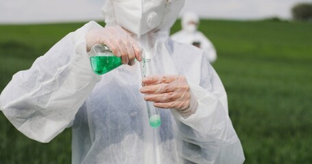 Caucasian woman scientist in protective clothing holding tube with pesticides liquid and doing experiment in field. Laboratory male and female workers working with chemical substance.