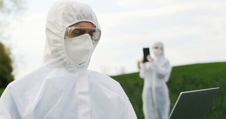Male Caucasian farmer biologist in white protective costume, mask and goggles standing in green...