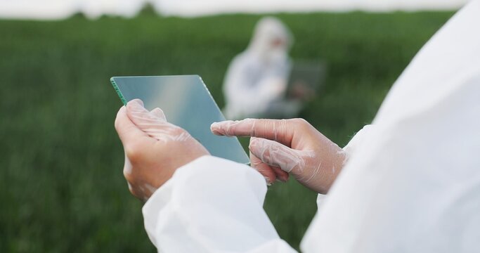 View over shoulder of farmer ecologist in protective suit tapping on glass transparent screen. Close up of futuristic device in field Touchscreen of hi-tech Scientist with tablet of future in hands.