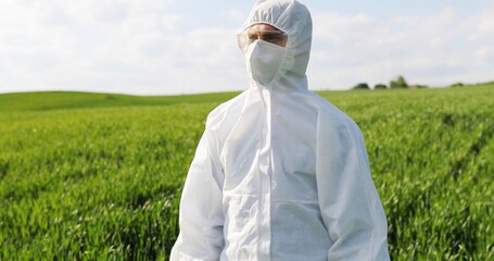 Portrait of Caucasian male farmer ecologist in white protective costume, mask and goggles standing in green field and turning face to camera. Man scientist and biologist posing in margin with harvest.