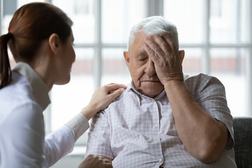 Female doctor therapist comforting older man feeling unwell during visit close up, touching head,...