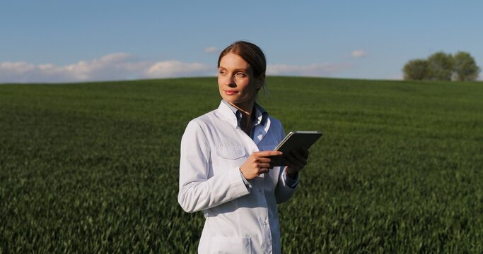 Caucasian beautiful female ecologist scientist in white gown walking in green field and holding tablet device. Woman researcher, biologist strolling in margin of harvest while using tablet computer.