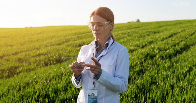Caucasian beautiful female ecologist scientist in white gown, mask and goggles standing in green field and working on tablet device. Woman researcher, biologist in margin using tablet computer.