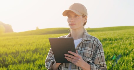 Portrait of beautiful young female farmer in hat standing in field and using tablet device. Pretty Caucasian woman tapping and scrolling on tablet computer in green margin with harvest in summer.