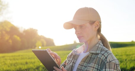 Portrait of attractive female farmer in hat holding tablet device and smiling to camera in green...
