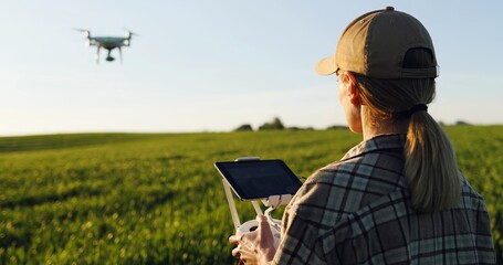 Close up of rear of Caucasian woman farmer in hat standing in green wheat field and controlling of drone which flying above margin. Female using tablet device as controller. Technologies in farming. - obrazy, fototapety, plakaty