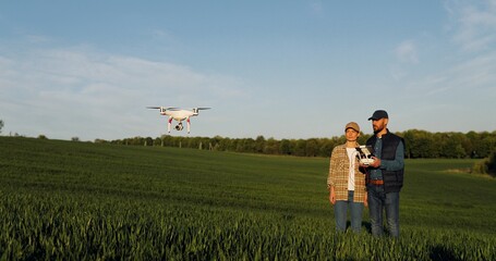 Caucasian happy couple of farmers standing in green wheat field and controlling of drone which flying above margin. Husband and wife using tablet device as controller. Technologies in agriculture.