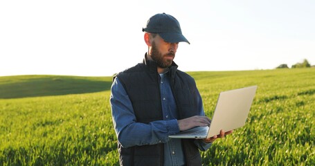 Caucasian young male farmer in hat standing in field and typing on keyboard of laptop computer....