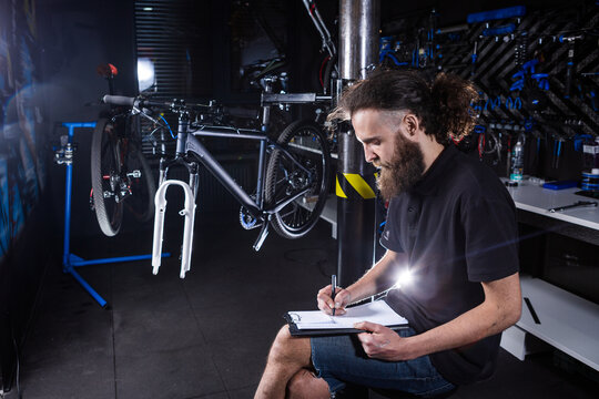 Stylish man with beard, cycle mechanic takes notes, checklist before repairing and servicing bike in bicycle shop workshop. Caucasian male in bicycle shop writing on clipboard. Assessing bike supply - Powered by Adobe