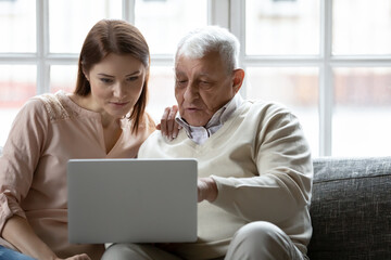 Young woman and older man using laptop together, looking at screen, sitting on couch at home,...