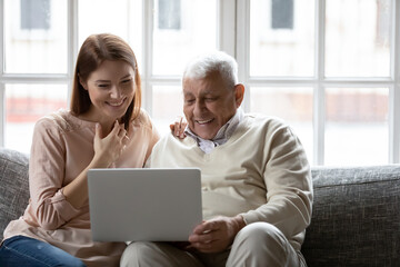 Happy older father and grownup daughter looking at laptop screen, having fun with computer, smiling mature man and young woman watching movie, shopping online, sitting on cozy couch at home