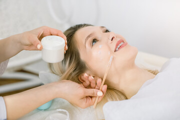 Naklejka premium Close up of female beautician hands, holding jar with cream, and applying it using wooden spatula to patient face in cosmetology clinic. Young girl having beauty procedures in spa