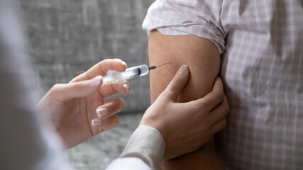 Close up young female nurse vaccinating older man, using syringe, touching arm, disease prevention...