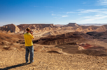 A young man on top of a mountain photographs a mountain landscape at the camera. Ramon Crater,...