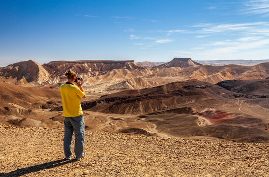 A young man on top of a mountain photographs a mountain landscape at the camera. Ramon Crater, Israel. Travel and photos concept.