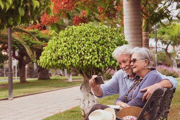 Couple of senior woman and man with white hair relaxing on a bench in public park taking a selfie - concept of active elderly during retirement