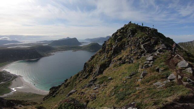 A group of hikers are walking up the trail to the peak of mountain Mannen, overlooking Haukland beach and the fjords in the background in Lofoten, Norway.